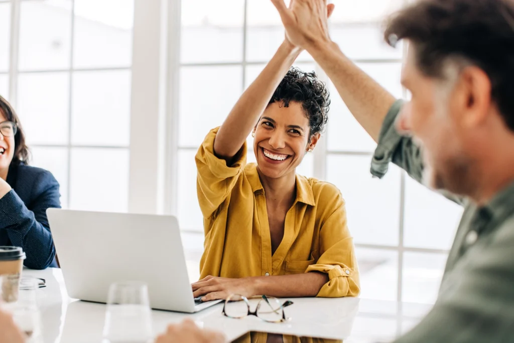 Country-Bank-coworkers-high-five-at-table-in-office-while-another-coworker-looks
