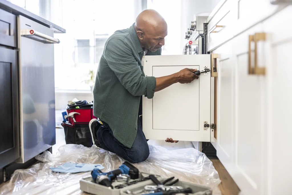 Country-Bank-man-kneeling-in-kitchen-fixing-cabinet-with-screwdriver