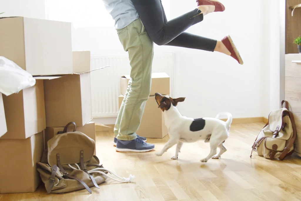 Country-Bank-man-lifting-woman-up-in-hug-while-dog-watches-and-boxes-are-piled-around-new-home